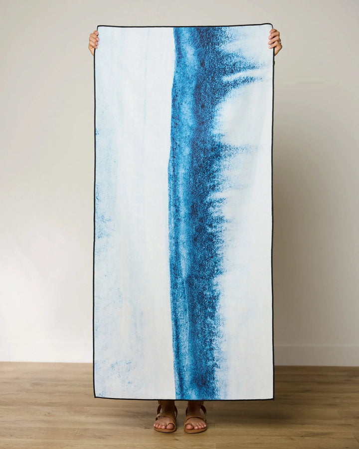 A person stands against a plain wall, holding up the GEOMETRY In The Distance quick-drying beach towel, featuring vertical blue abstract stripes on white. Only their feet and fingers are visible.