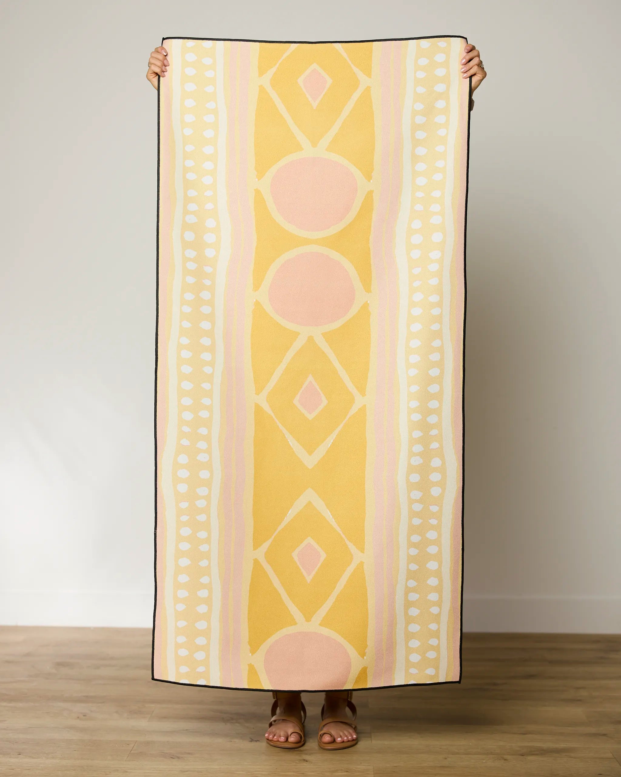 A person stands indoors holding up the GEOMETRY Beaded quick-drying beach towel in yellow, peach, and white geometric patterns, with only their hands and sandaled feet visible behind it.