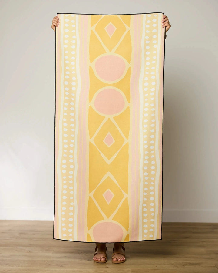 A person stands indoors holding up the GEOMETRY Beaded quick-drying beach towel in yellow, peach, and white geometric patterns, with only their hands and sandaled feet visible behind it.