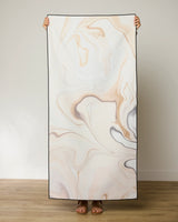 A person stands on a wooden floor holding up a rectangular GEOMETRY Choco Beach Towel; only their hands and feet are visible.