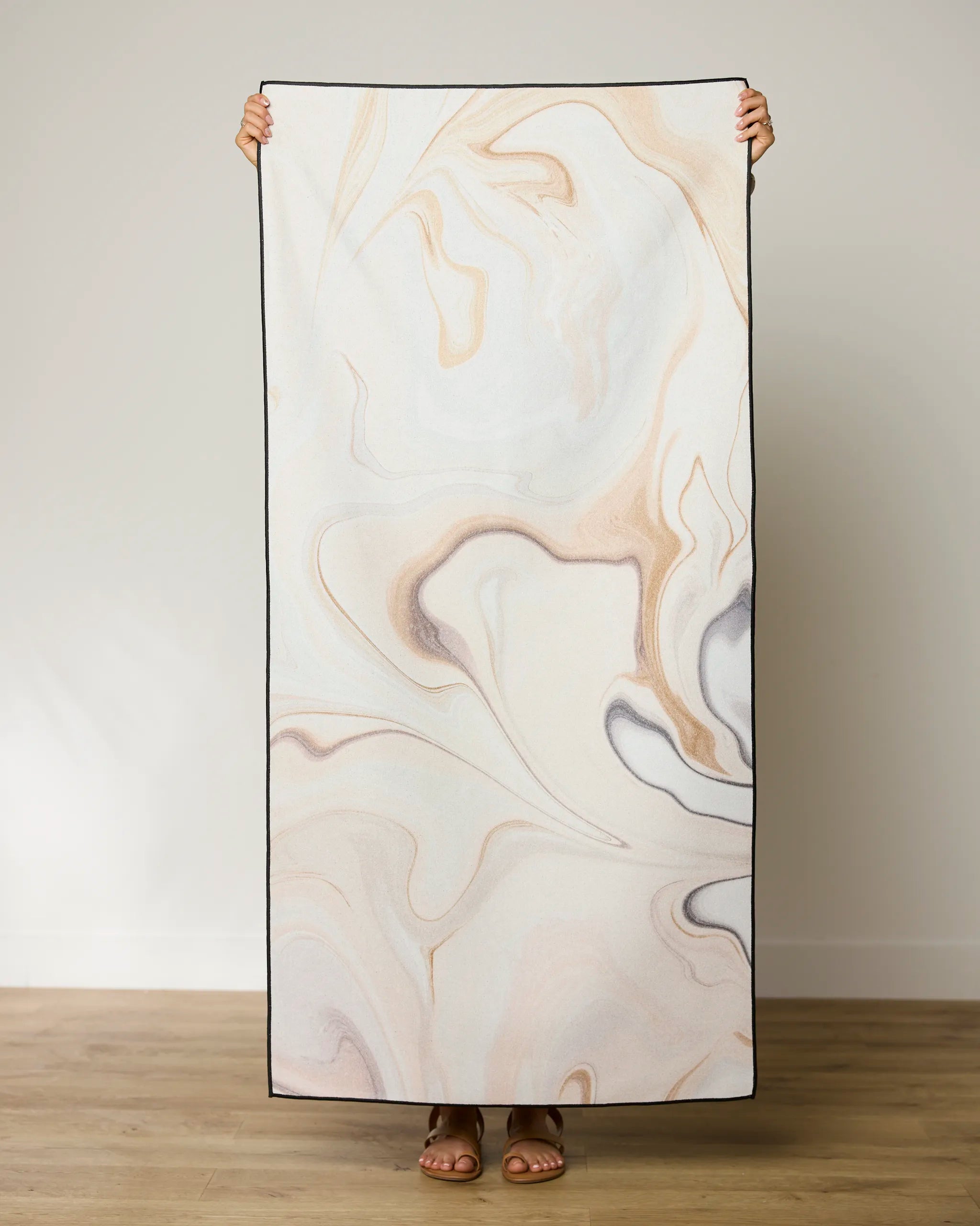 A person stands on a wooden floor holding up a rectangular GEOMETRY Choco Beach Towel; only their hands and feet are visible.