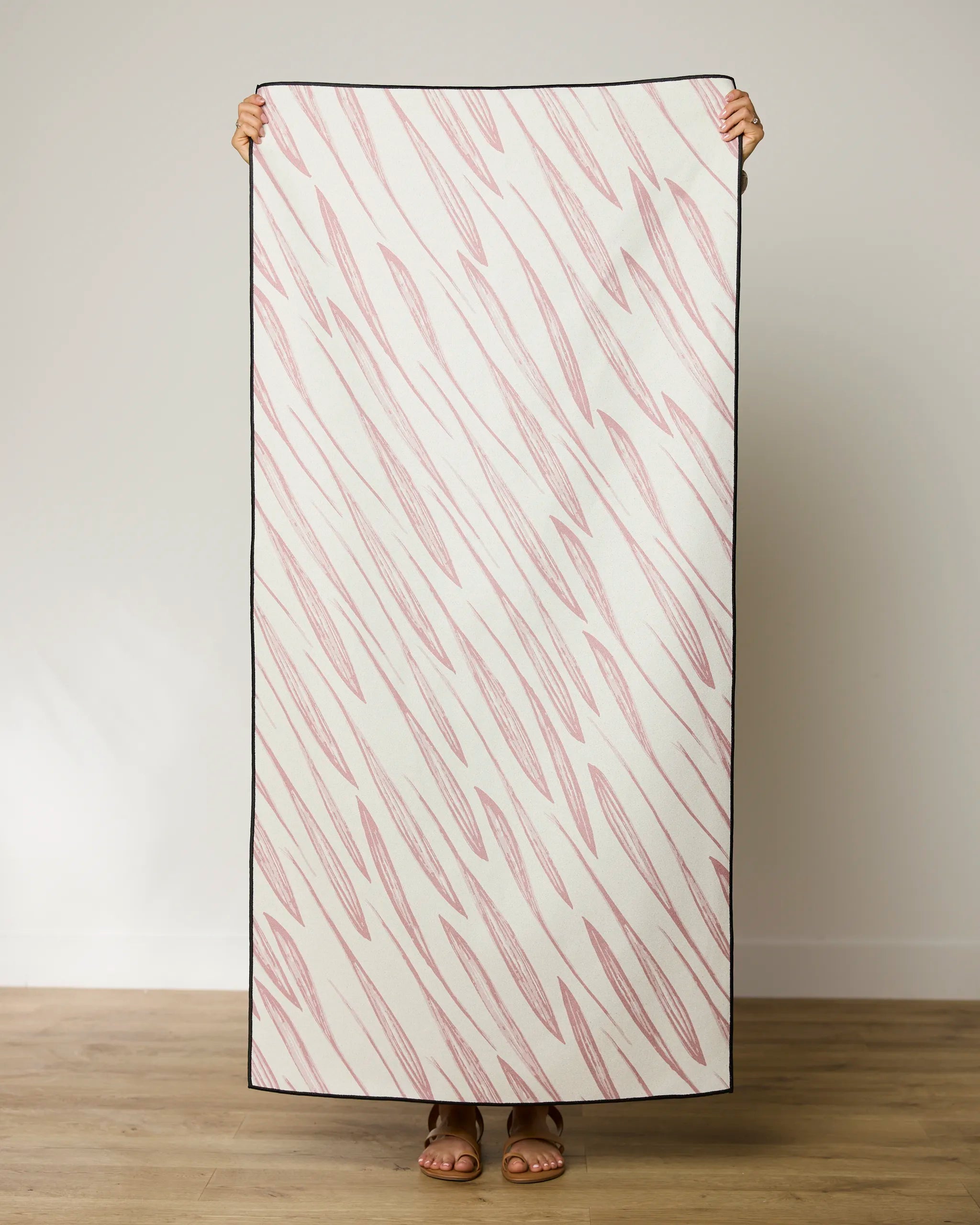 A person holds up the GEOMETRY Calm Winds Beach Towel, a rectangular quick-drying towel made from recycled materials, featuring a white background with diagonal pink zigzag lines. Only their hands and feet are visible behind the towel.