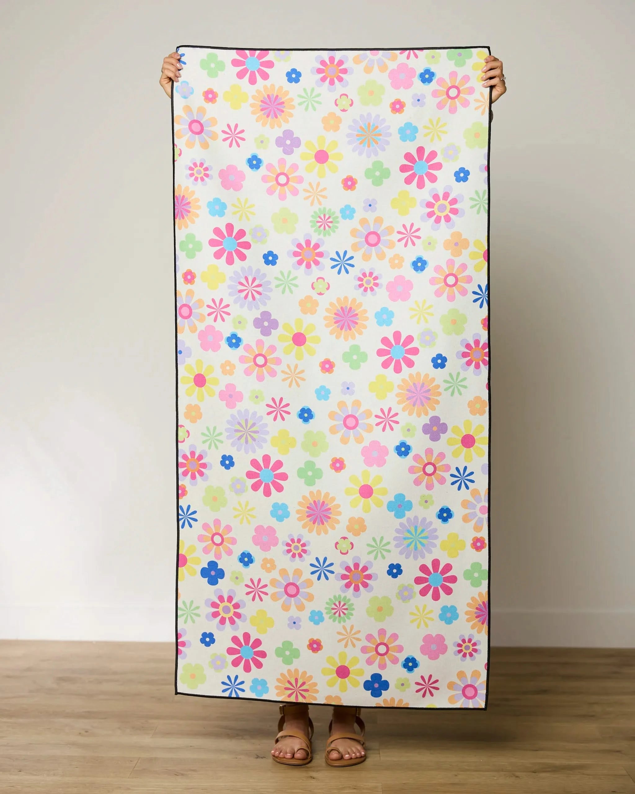 A person stands indoors holding up the GEOMETRY Far Out Flowers quick-drying beach towel, which features a colorful floral pattern with pink, yellow, blue, and purple flowers on a light background. Their face is hidden behind the towel.