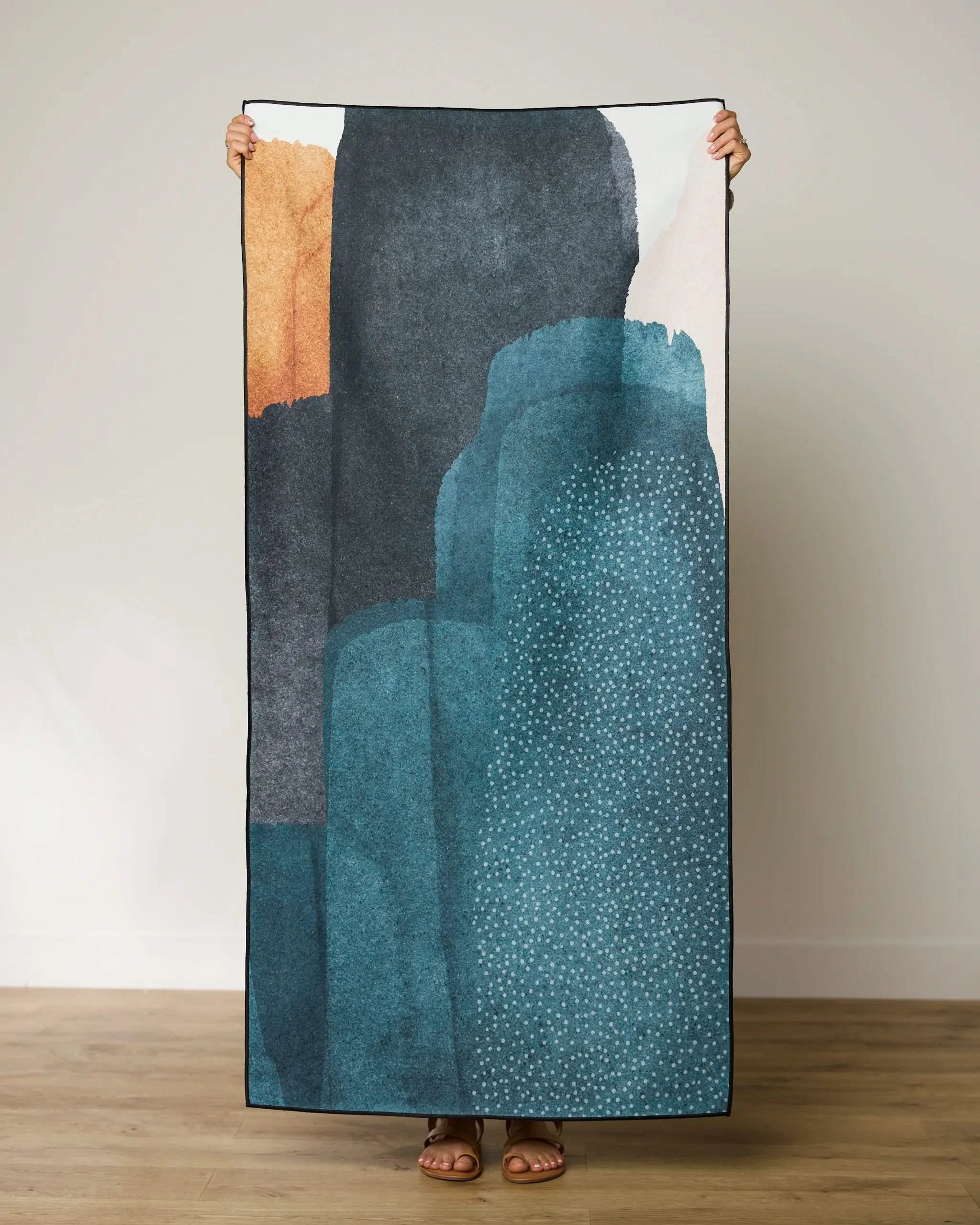 A person stands on a wooden floor holding up the GEOMETRY Founded Beach Towel, featuring an abstract design in teal, navy, orange, and white shapes that partially obscure their body.