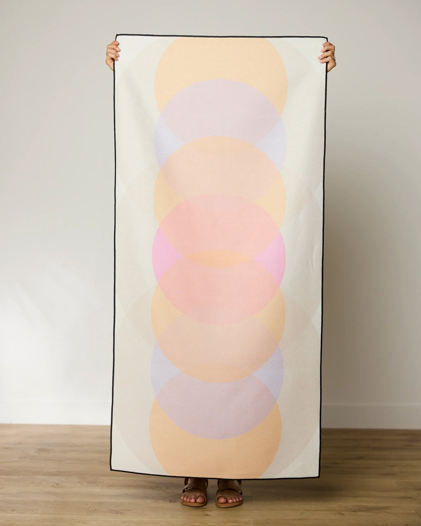 A person stands against a plain wall holding a GEOMETRY Funky Phases Beach Towel featuring overlapping pastel circles in peach, pink, and lavender. Only their hands, feet, and sandals are visible.