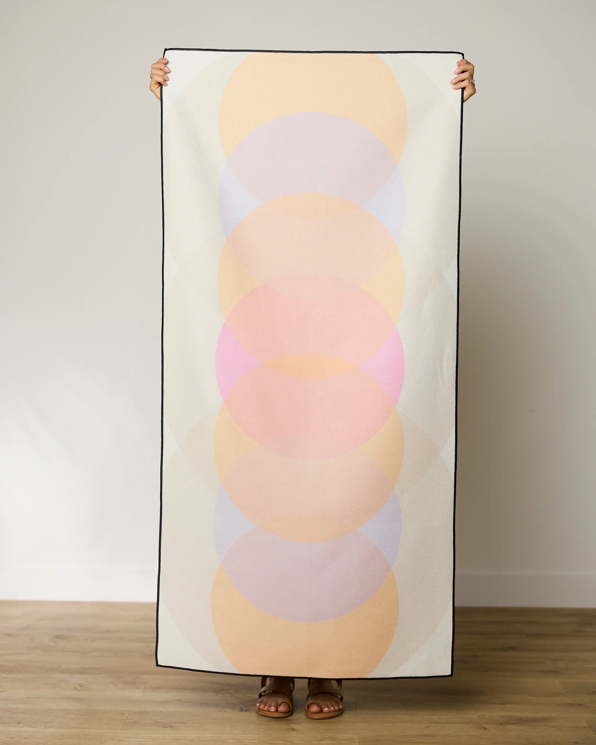 A person stands against a plain wall holding a GEOMETRY Funky Phases Beach Towel featuring overlapping pastel circles in peach, pink, and lavender. Only their hands, feet, and sandals are visible.