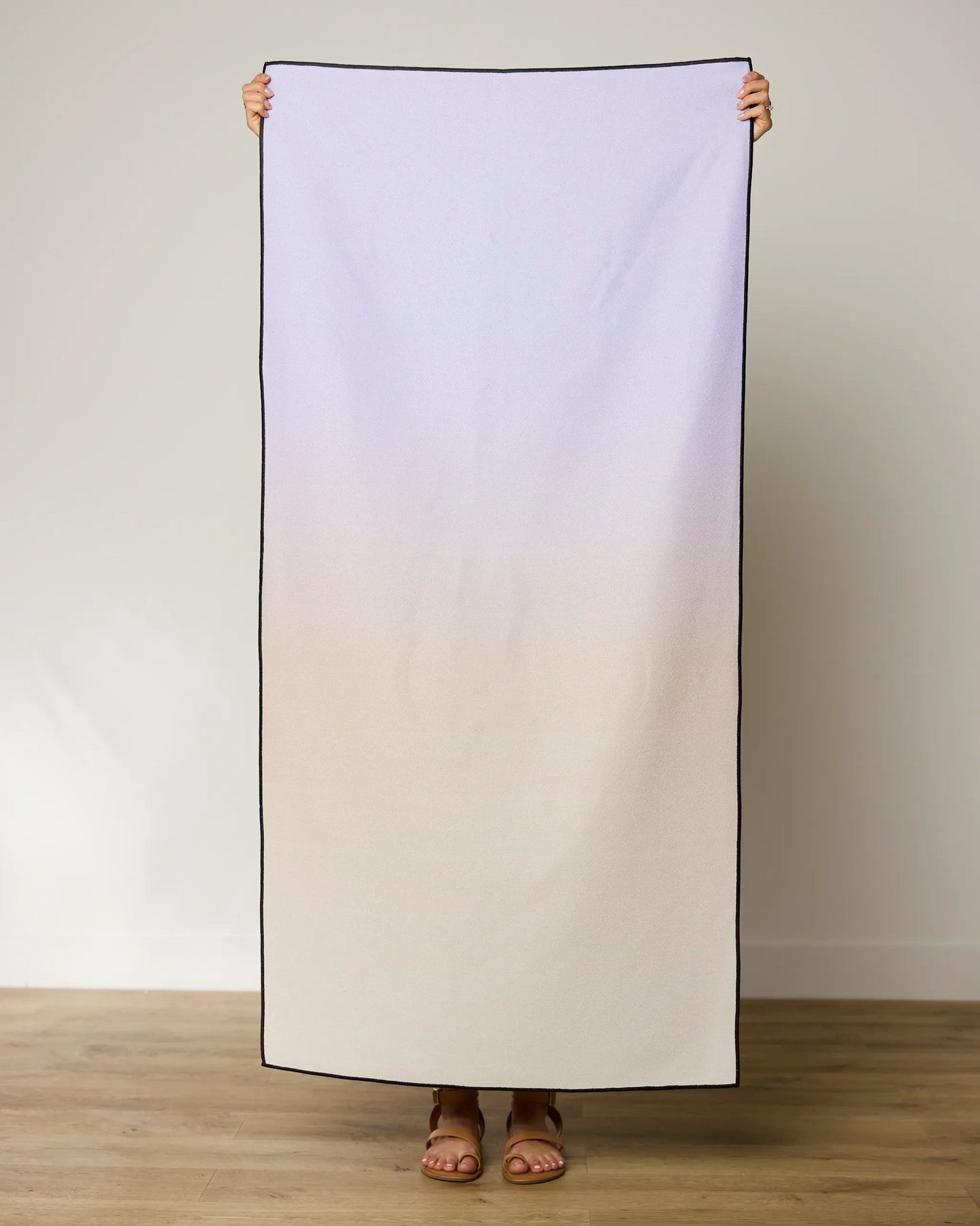 A person stands on a wooden floor holding up the GEOMETRY Groovy Gradient Micro-loop Beach Towel, showcasing its pale purple to beige gradient with black borders. Their feet in sandals peek out below this quick-drying towel made from recycled materials.