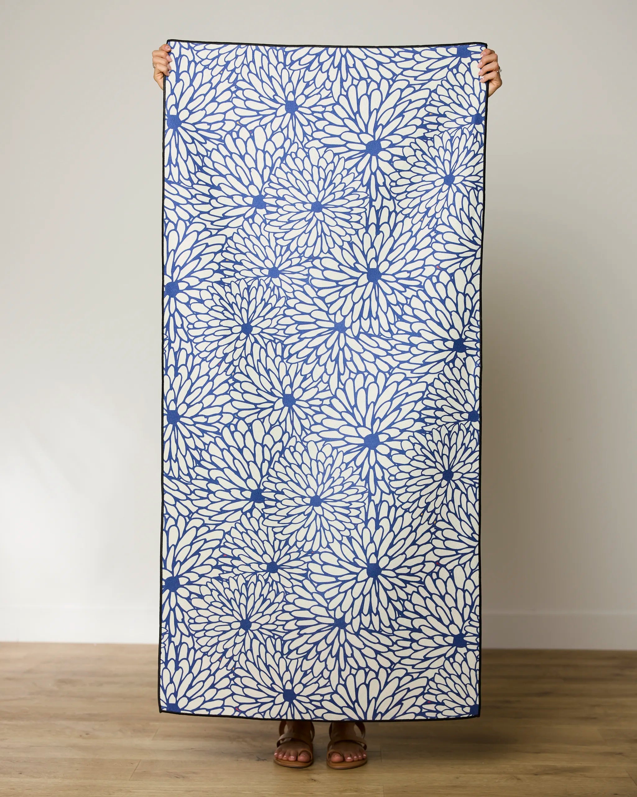 A person holds up a rectangular, blue and white floral GEOMETRY Jar Jar Micro-loop Beach Towel indoors on a wooden floor against a plain light wall. Only their hands, feet, and sandals are visible.