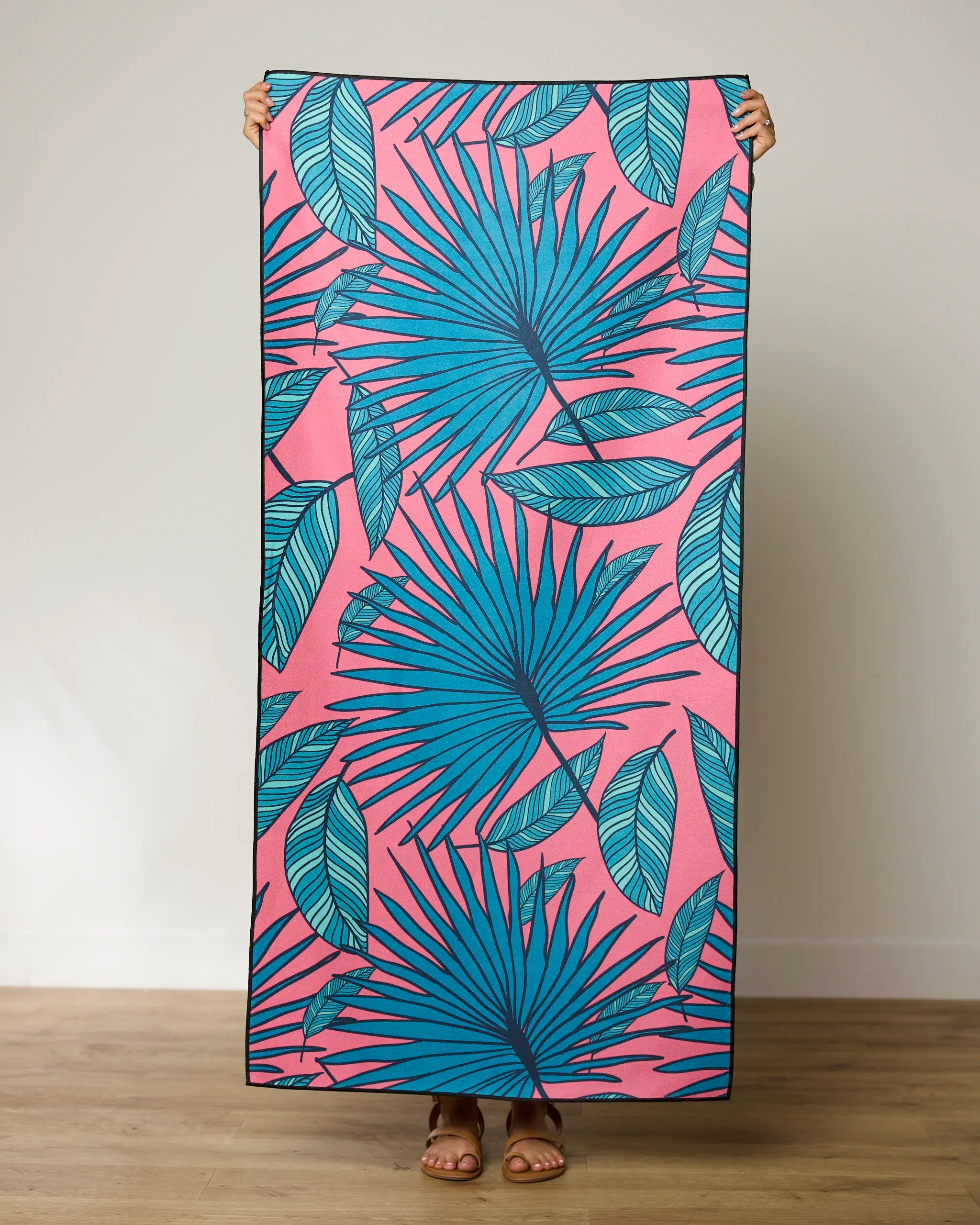 A person holds the GEOMETRY Jazzy Jungle micro-loop beach towel, featuring a pink background with bold blue tropical leaves. Only their hands and feet are visible indoors on a wooden floor. This quick-drying towel adds stylish flair anywhere.