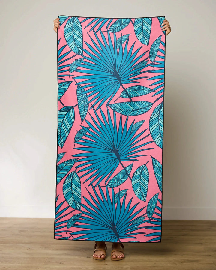 A person holds the GEOMETRY Jazzy Jungle micro-loop beach towel, featuring a pink background with bold blue tropical leaves. Only their hands and feet are visible indoors on a wooden floor. This quick-drying towel adds stylish flair anywhere.