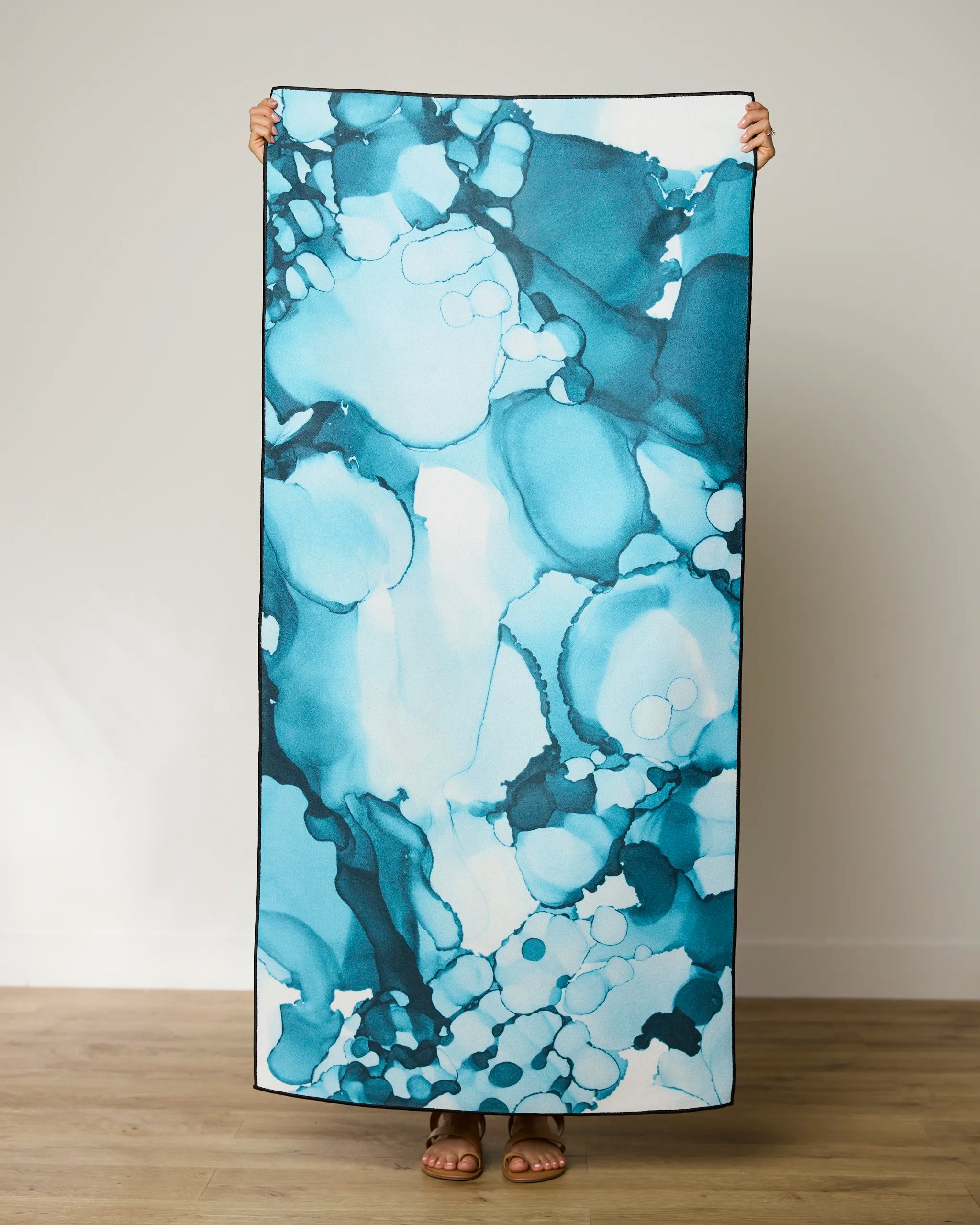 A person stands indoors holding the GEOMETRY Leah beach towel, featuring a large, quick-drying design with a blue and teal abstract pattern, covering their body except for their sandal-clad feet.