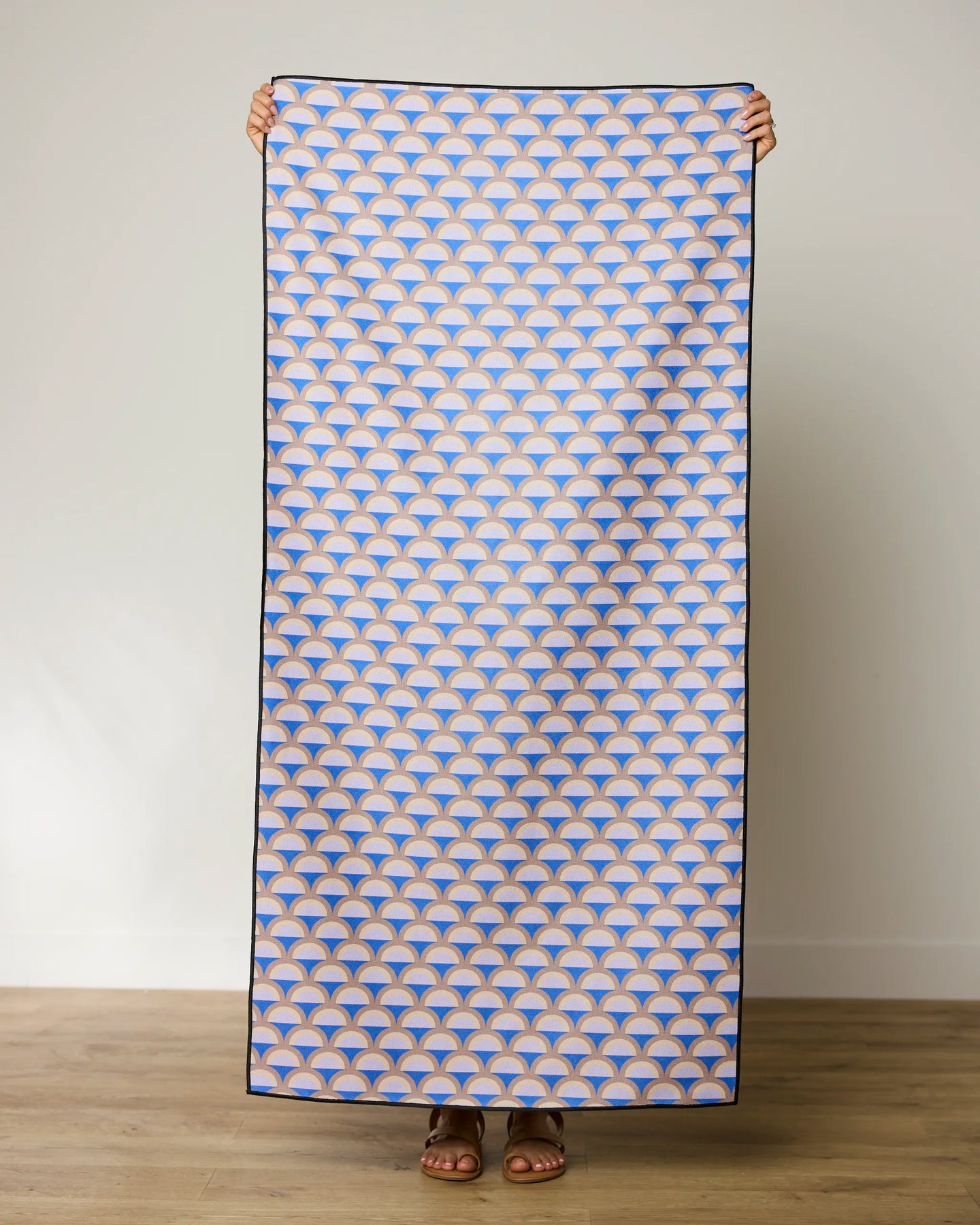 A person stands holding up the GEOMETRY Mauve Mounds quick-drying beach towel, which features a repeating blue, orange, and white geometric fan pattern, against a plain light wall and wooden floor.