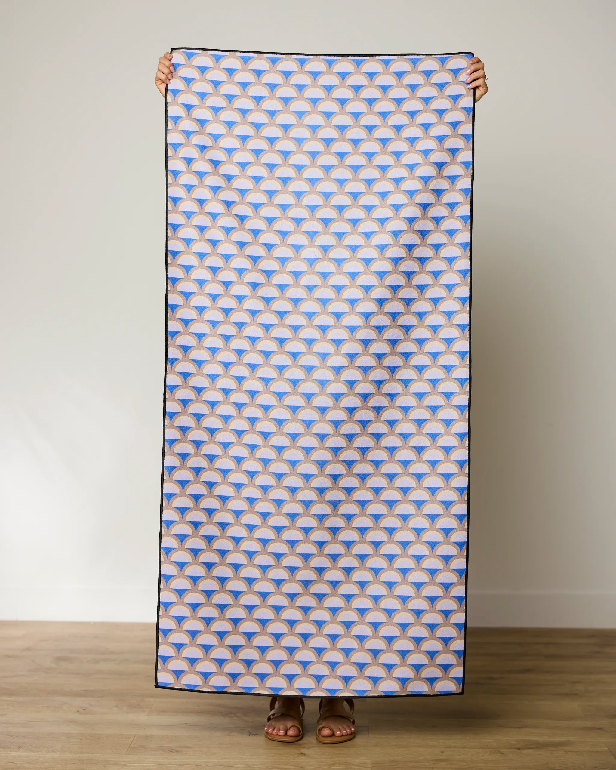 A person stands holding up the GEOMETRY Mauve Mounds quick-drying beach towel, which features a repeating blue, orange, and white geometric fan pattern, against a plain light wall and wooden floor.