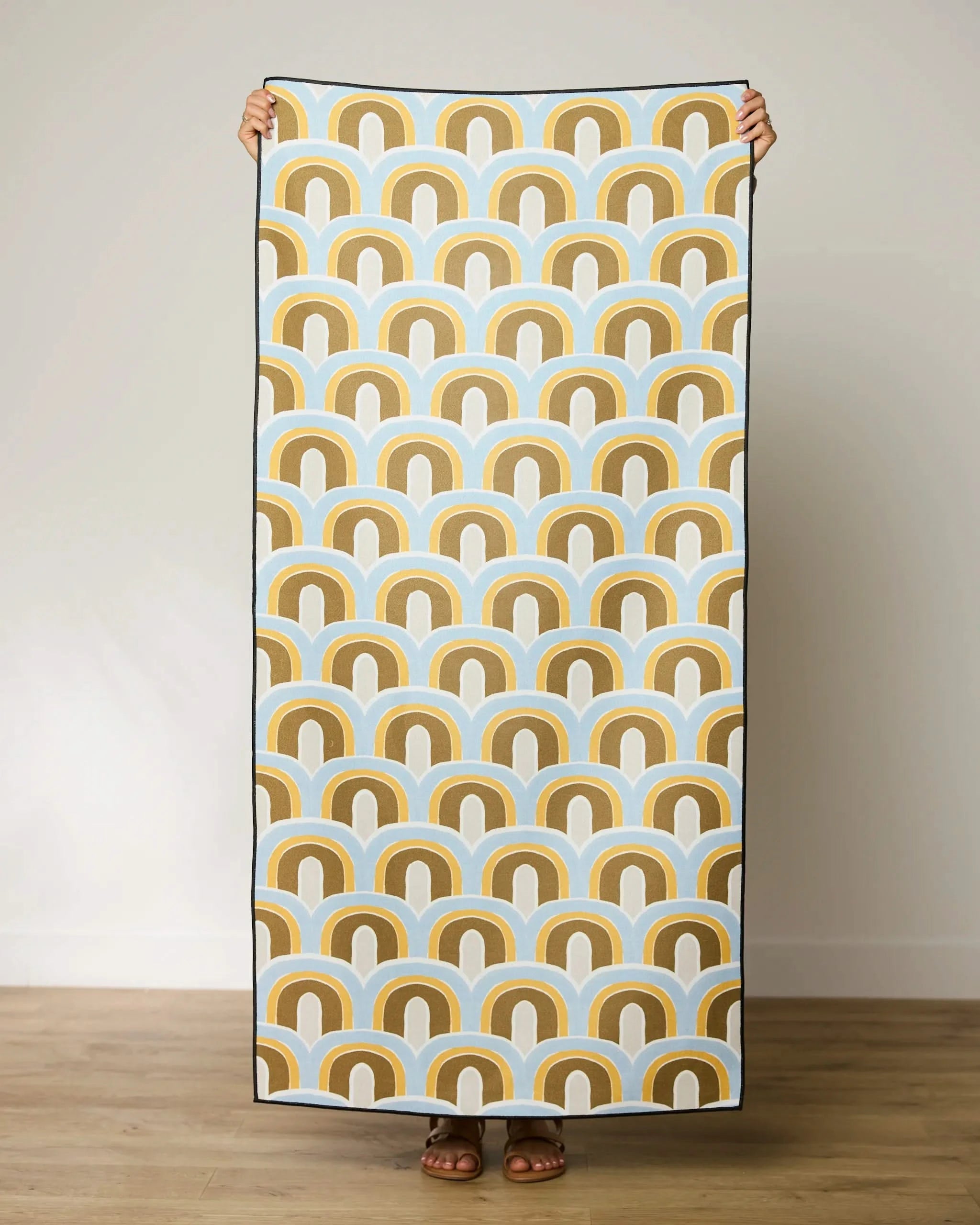 A person stands against a plain wall holding up the GEOMETRY Rained On Micro-loop Beach Towel, featuring a retro pattern of brown, mustard yellow, white, and blue arches. Only their hands and feet are visible. Made from recycled materials.