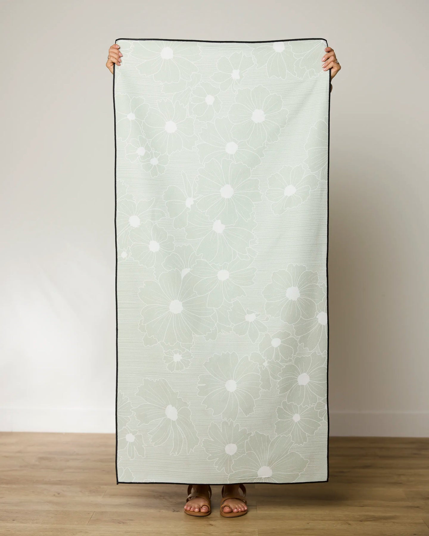 A person stands indoors holding up the GEOMETRY Sage Petals Beach Towel, which features a light green background with a white floral pattern. Made from recycled materials, the quick-drying towel covers their face and body.