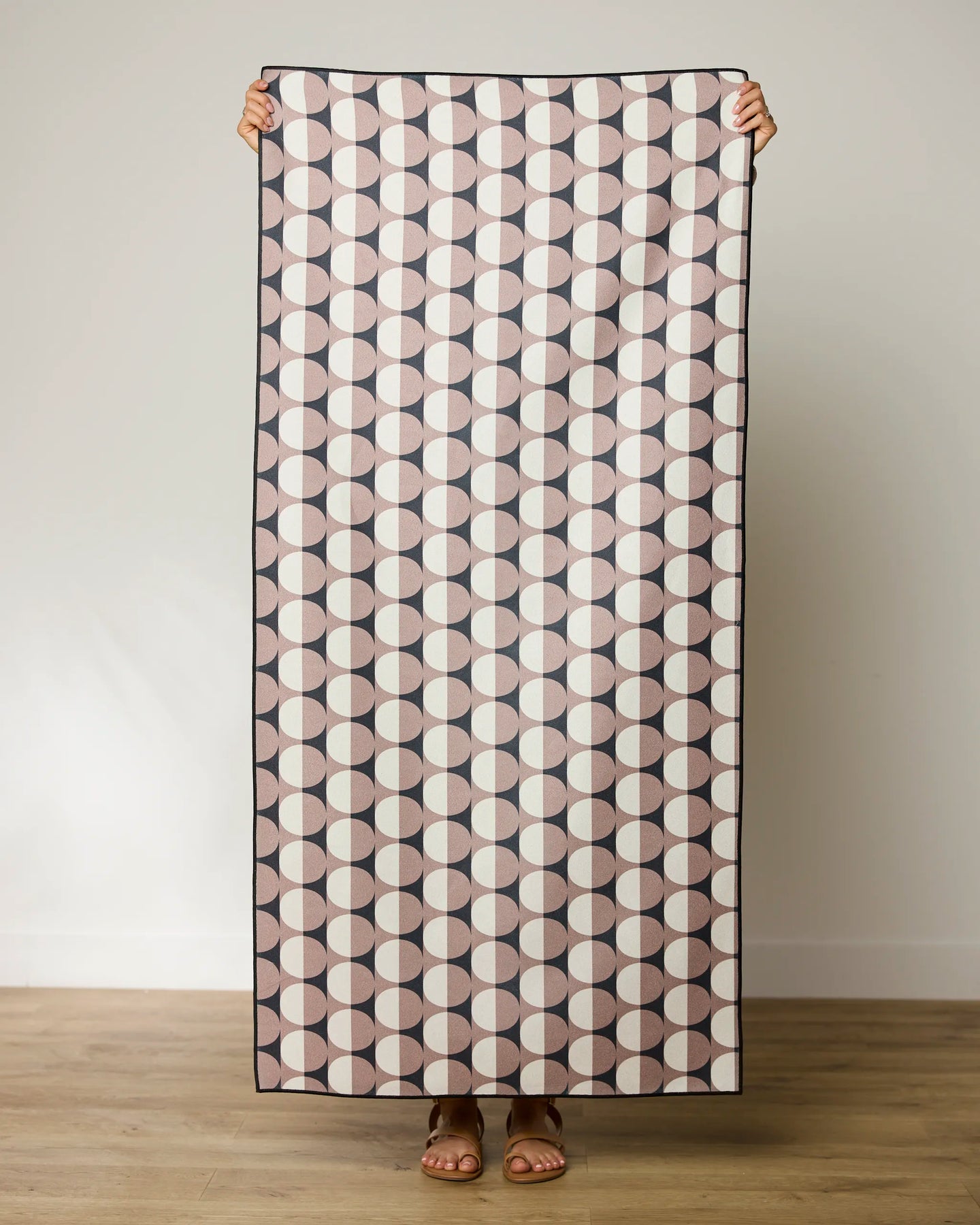 A person holds up a GEOMETRY Retro Round Light towel, featuring a repeating pattern of circles in pink, cream, and dark brown halves. Only their hands, feet, and sandals are visible against a plain wall and wooden floor.