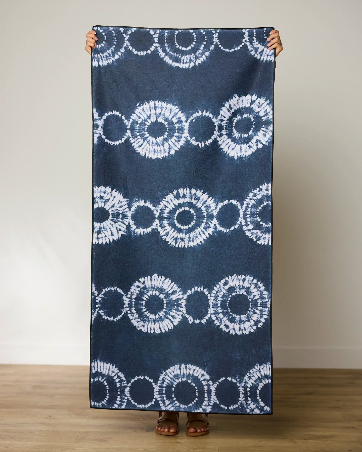 A person stands on a wooden floor holding up the GEOMETRY Shibo Stars large blue and white micro-loop beach towel made from recycled materials, covering their body except hands and feet, against a plain light wall.