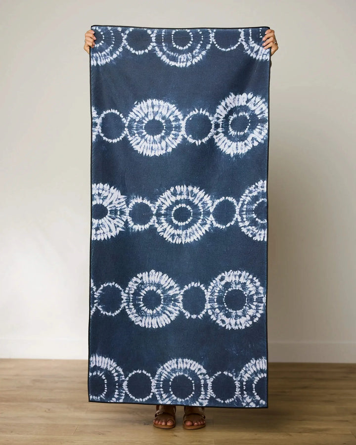 A person stands on a wooden floor holding up the GEOMETRY Shibo Stars large blue and white micro-loop beach towel made from recycled materials, covering their body except hands and feet, against a plain light wall.