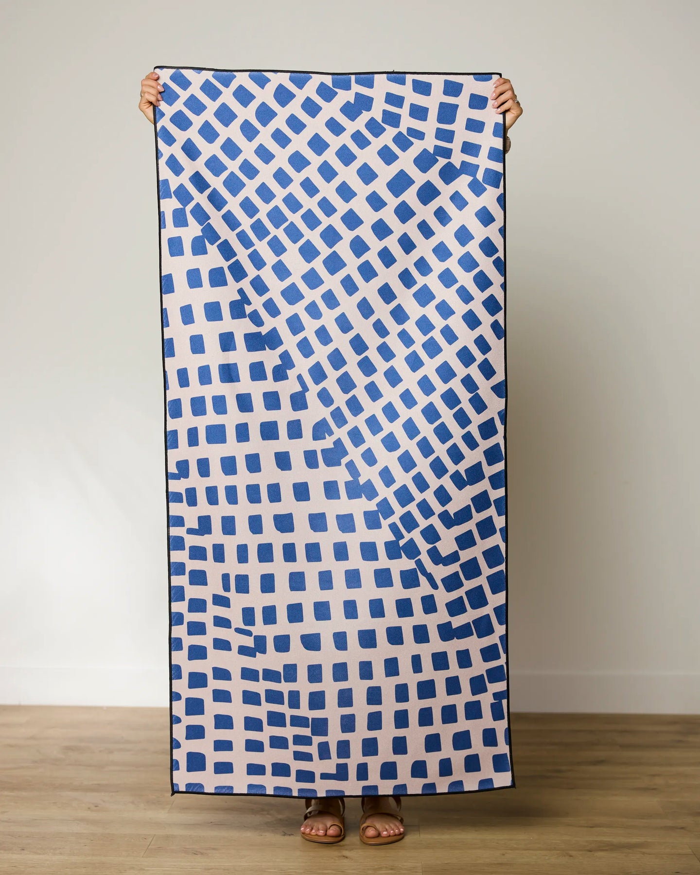 A person holds up the GEOMETRY Shelly Back towel, which displays irregular blue squares on a light background. Only their hands and feet are visible against a plain wall and wooden floor.