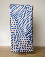 A person holds up the GEOMETRY Shelly Back towel, which displays irregular blue squares on a light background. Only their hands and feet are visible against a plain wall and wooden floor.