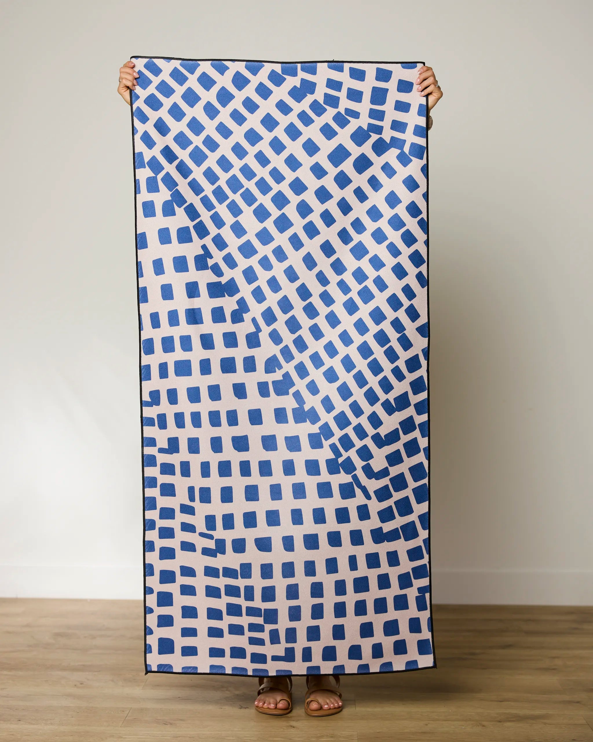 A person holds up the GEOMETRY Shelly Back towel, which displays irregular blue squares on a light background. Only their hands and feet are visible against a plain wall and wooden floor.