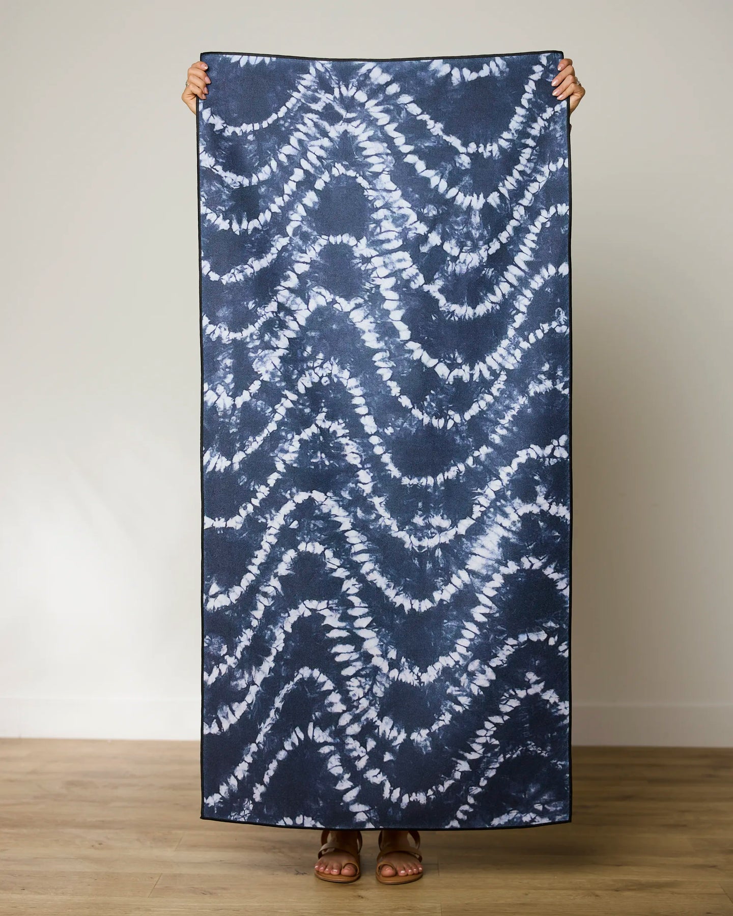 A person stands indoors holding up the rectangular Shibo Waves beach towel by GEOMETRY—navy blue and white, made from recycled materials—which covers their body except for hands, feet, and sandals.
