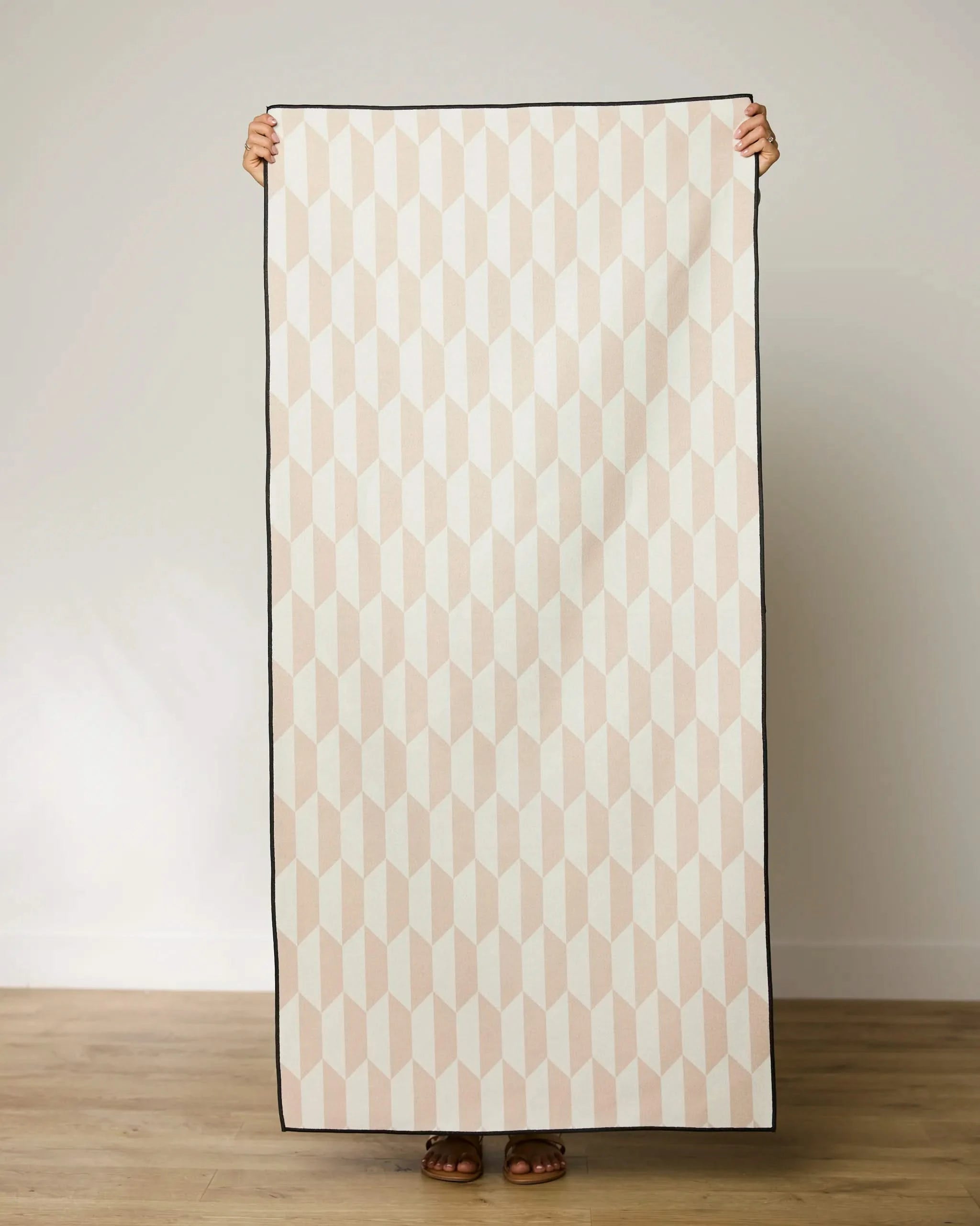 A person standing on a light wooden floor holds up a GEOMETRY Tan Trapezoid Micro-loop Beach Towel with a beige geometric pattern; only their feet and hands are visible.