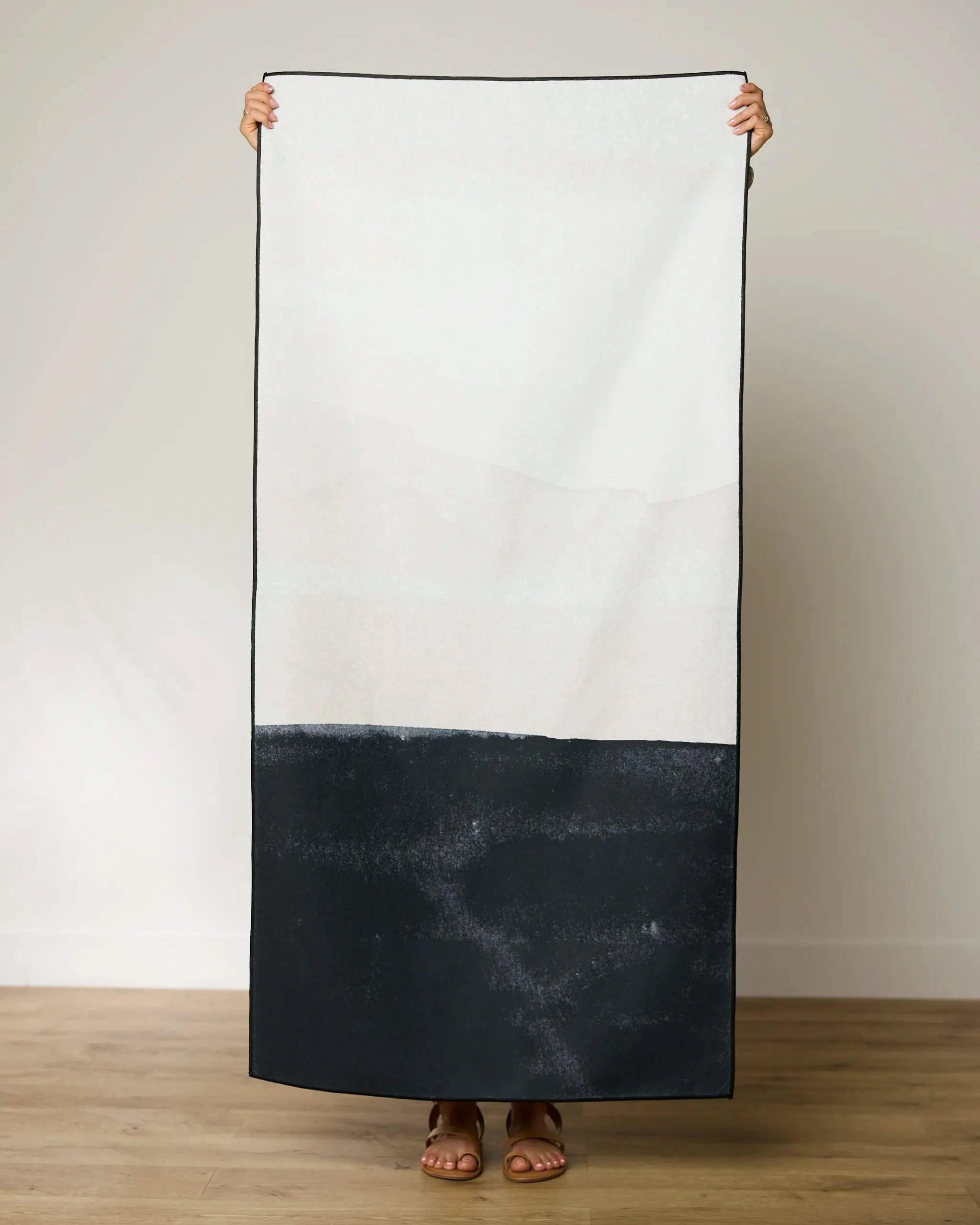 A person stands on a wooden floor, holding up a GEOMETRY Top Layer beach towel with an abstract black and white design made from recycled materials, their face hidden behind the quick-drying towel against a light wall.