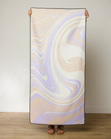 A person stands against a plain wall, holding up the GEOMETRY Way Out Wave towel in a pastel marble swirl of beige, light purple, and white; only their hands and feet are visible.