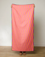 A person stands indoors holding up the GEOMETRY Bright Trapezoid beach towel, featuring a pink, yellow, and orange geometric pattern; only their hands, feet, and sandals are visible.