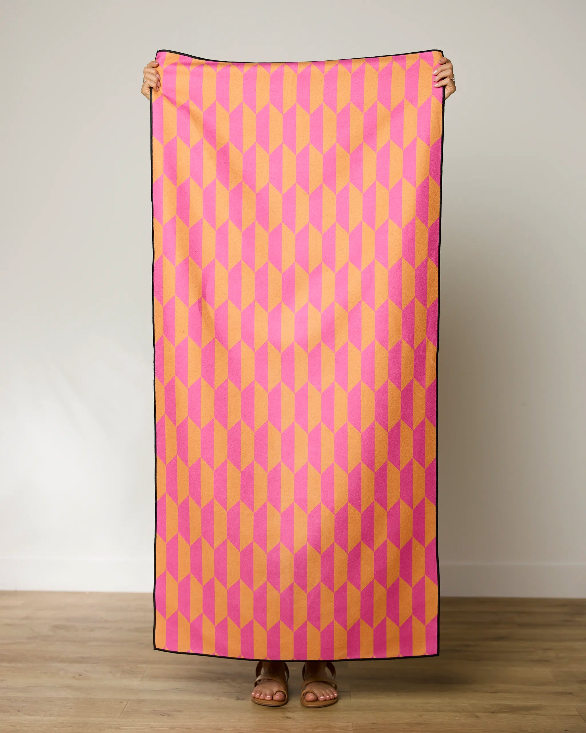 A person stands indoors holding up the GEOMETRY Bright Trapezoid beach towel, featuring a pink, yellow, and orange geometric pattern; only their hands, feet, and sandals are visible.