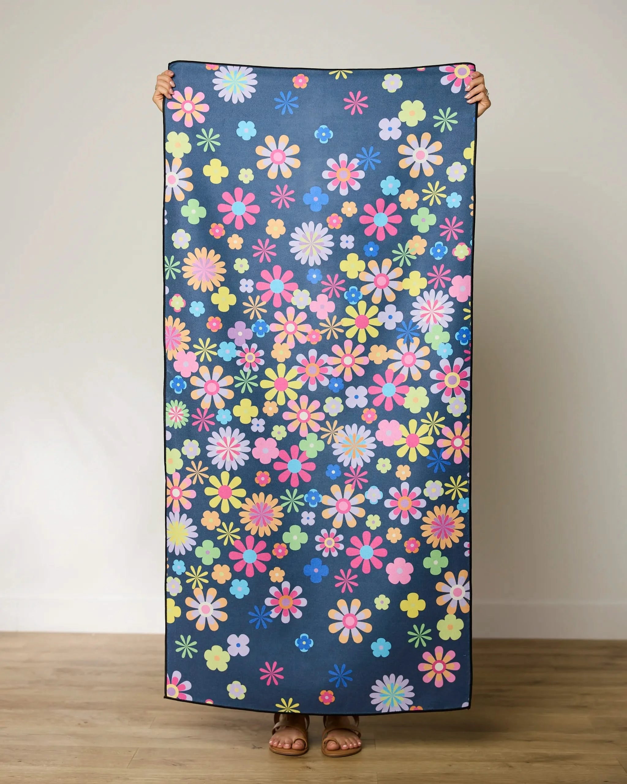 Indoors on a wooden floor, a person holds up the GEOMETRY Far Out Flowers Dark Micro-loop Beach Towel with a colorful floral pattern—pink, yellow, blue, and green flowers on dark blue. The quick-drying towel covers their face.