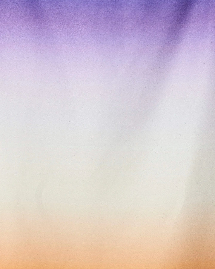 A soft gradient background transitions from purple to white to orange, evoking the Radiant quick-drying micro-loop beach towel by GEOMETRY, crafted from recycled materials with a smooth, calming color flow.