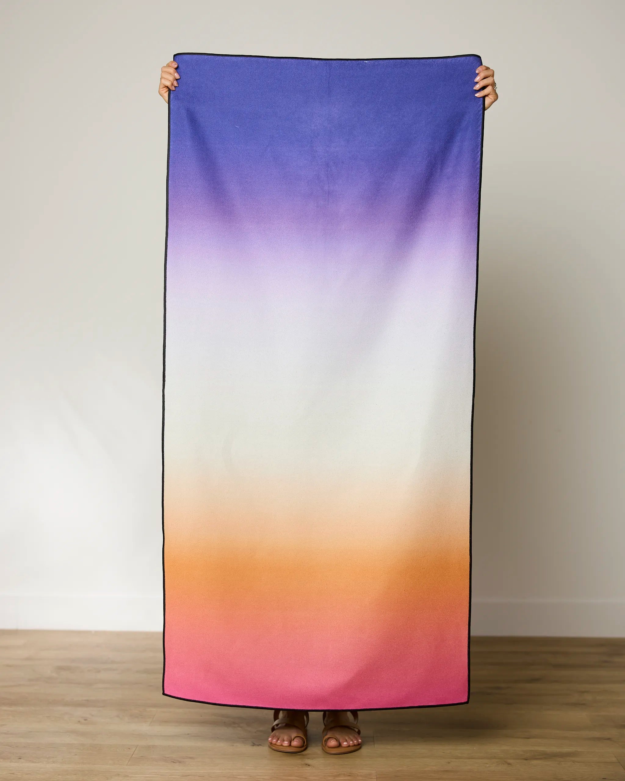 A person stands indoors on a wooden floor holding up the GEOMETRY Radiant micro-loop beach towel, featuring a blue-to-purple, white, orange, and pink gradient. Only their feet and hands are visible.