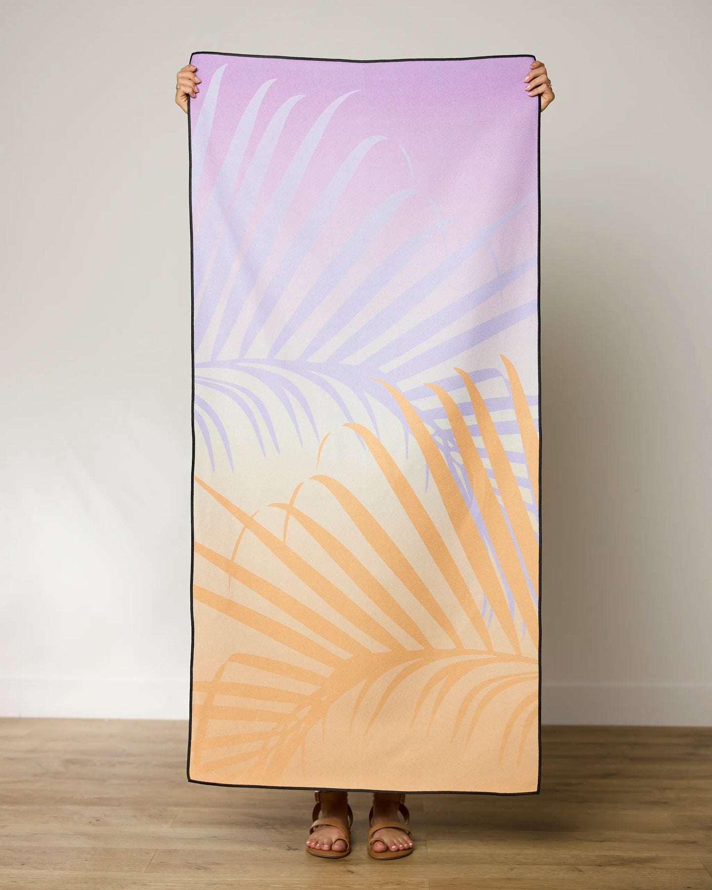 A person holds up the GEOMETRY Sunset Palm Micro-loop Beach Towel, featuring a pastel gradient design with overlapping purple and beige palm leaves, indoors on a wooden floor against a plain light wall.