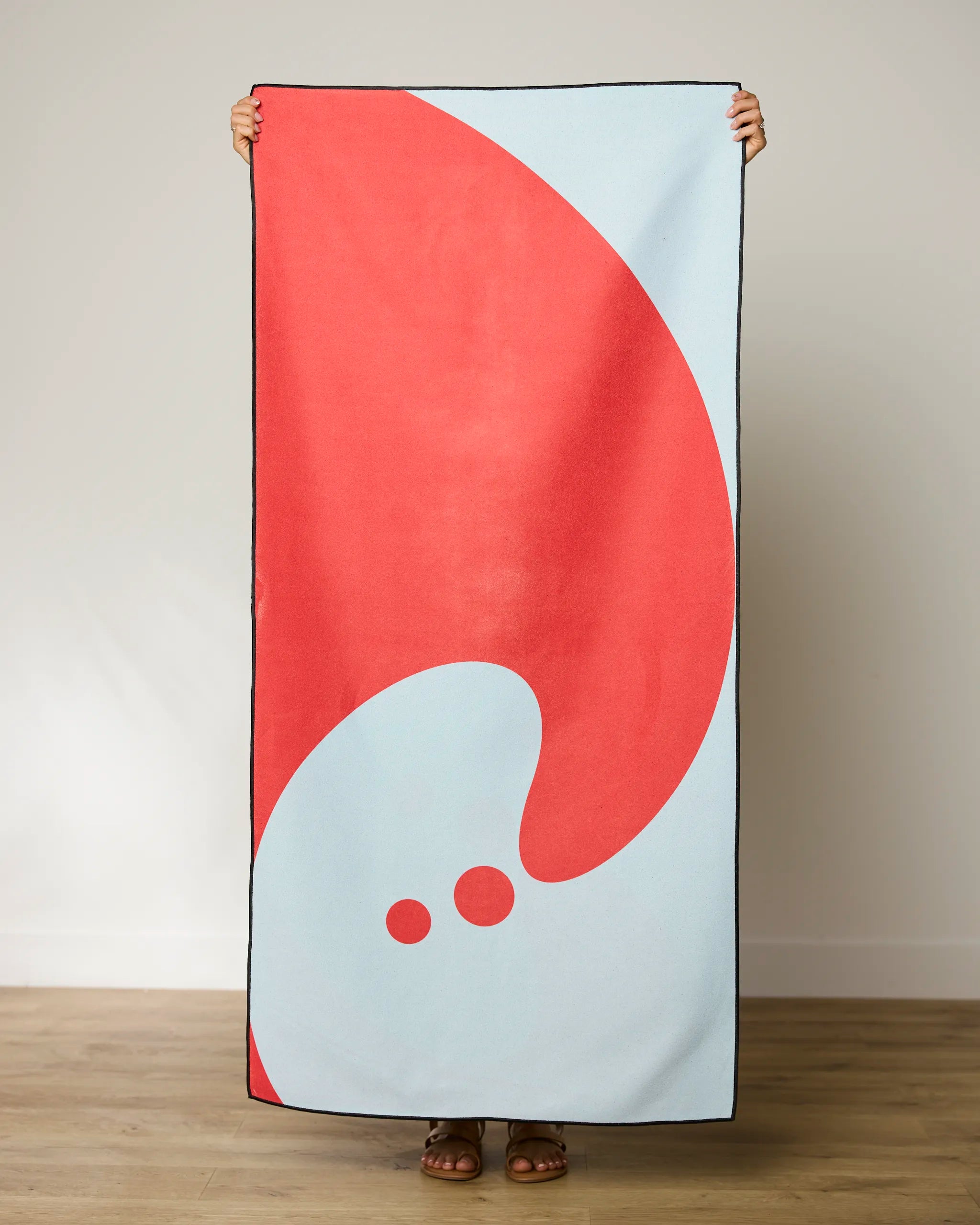 A person stands indoors on a wooden floor, holding the GEOMETRY Tidal Micro-loop Beach Towel—an abstract red and light blue design made from recycled materials—that covers their body, showing only their feet in sandals.