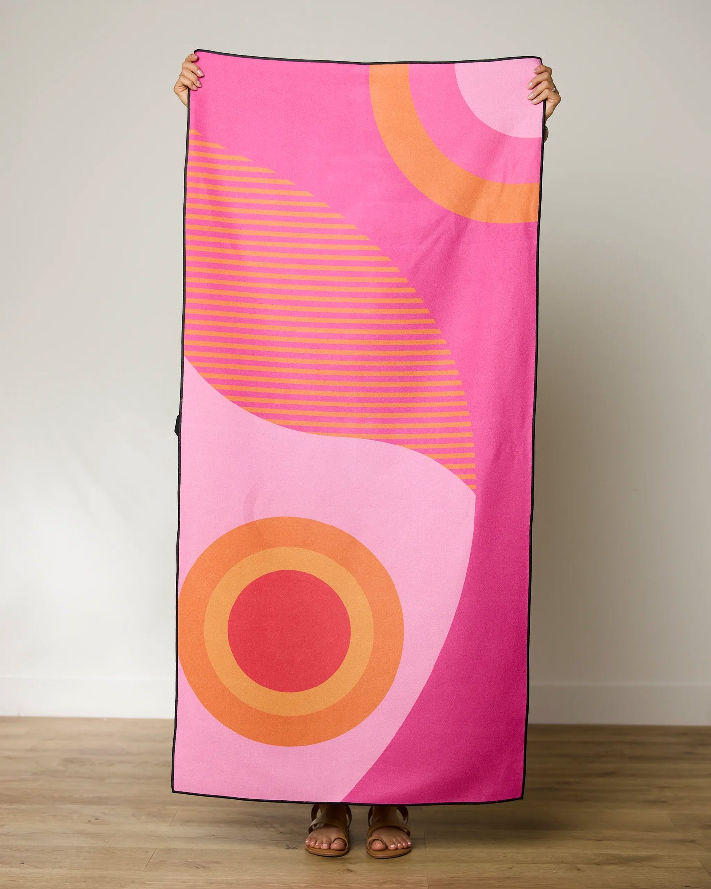 A person holds up the GEOMETRY Round Trip Micro-loop Beach Towel, featuring bold pink, orange, and red abstract patterns with curved lines and circles. Made from recycled materials, this quick-drying towel hides the persons face.