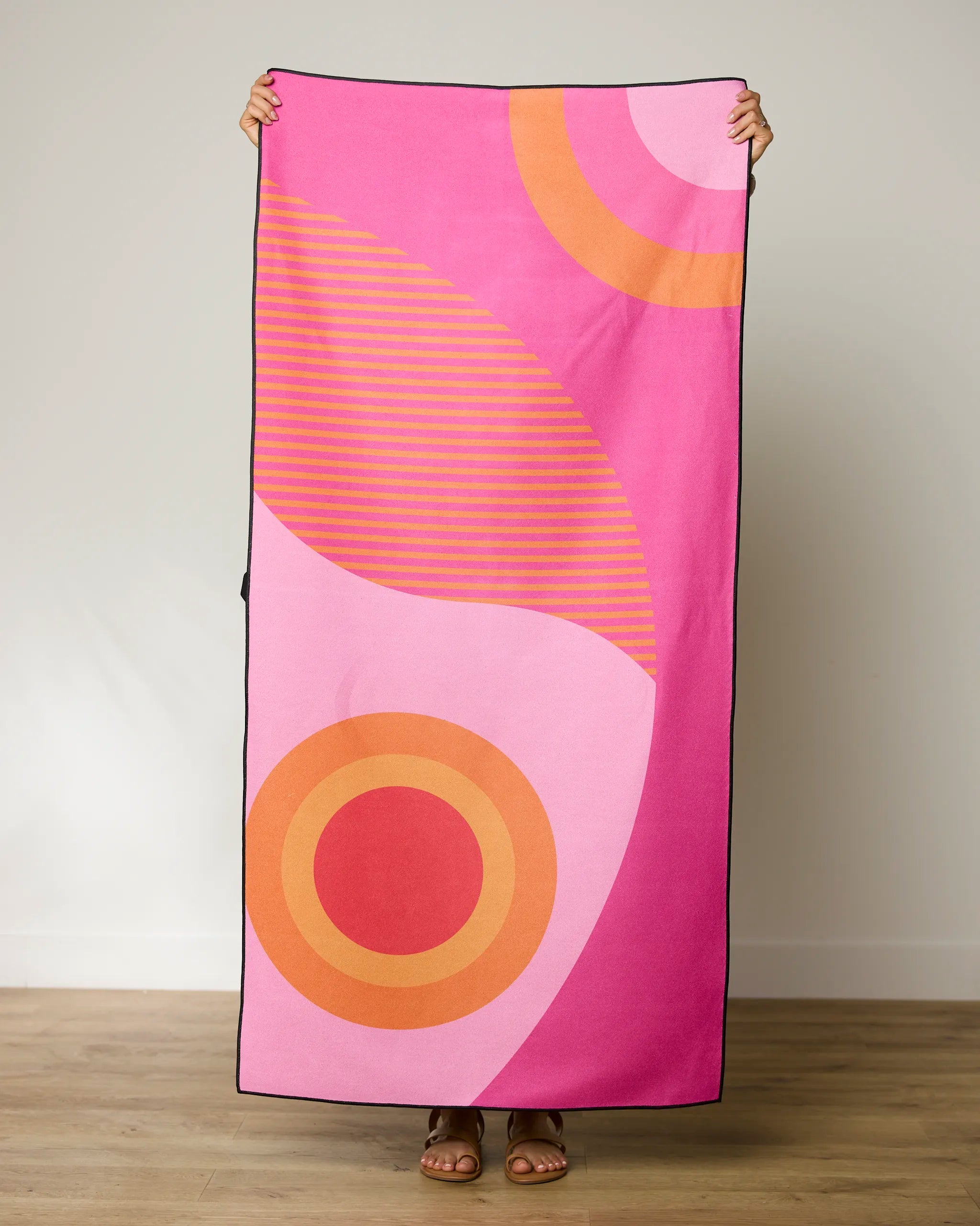 A person holds up the GEOMETRY Round Trip Micro-loop Beach Towel, featuring bold pink, orange, and red abstract patterns with curved lines and circles. Made from recycled materials, this quick-drying towel hides the persons face.