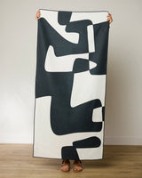 A person stands on a wooden floor, holding up a GEOMETRY Abstract Neutral Beach Towel with a black and white abstract pattern that obscures their face and body behind this quick-drying, stylish towel.