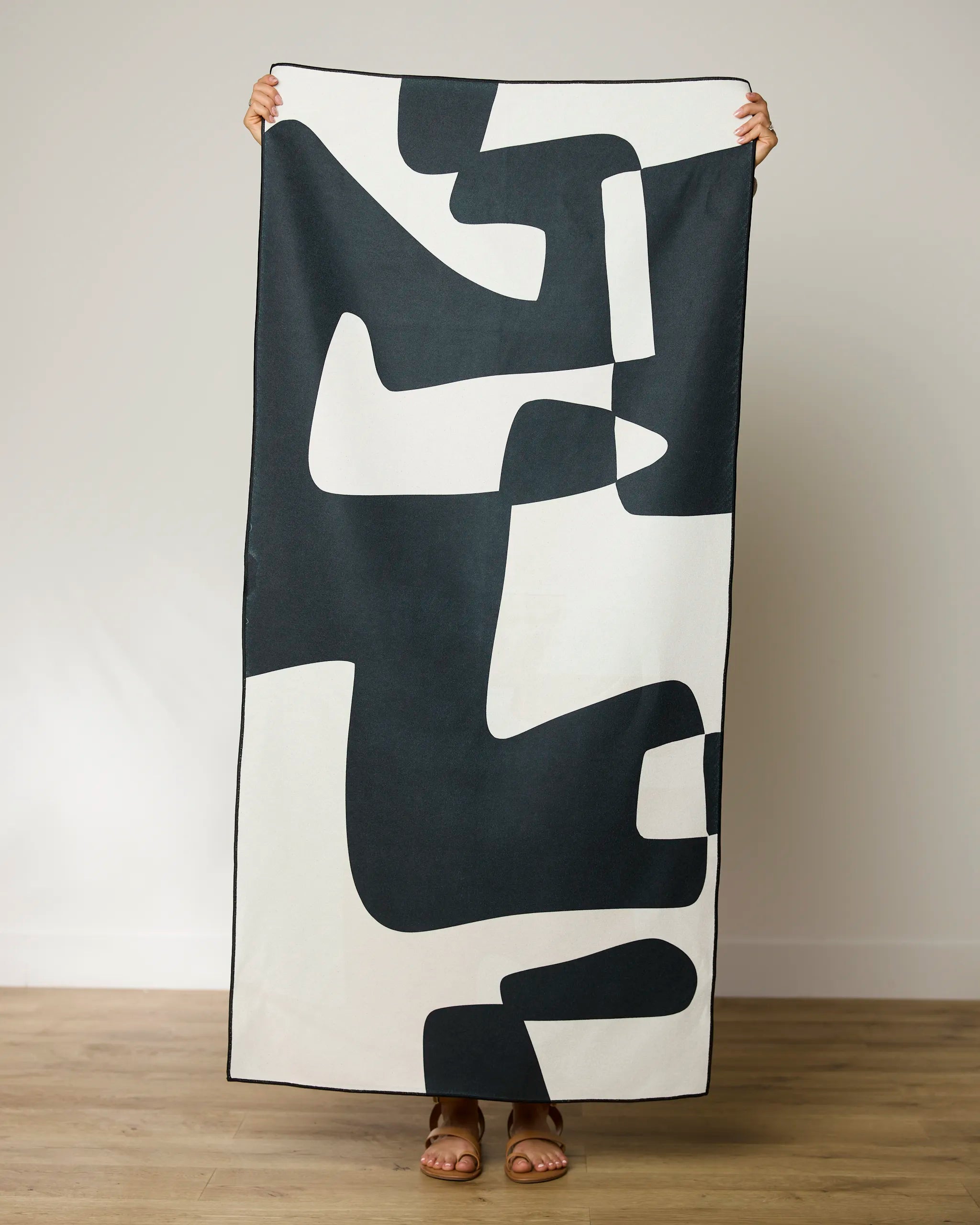 A person stands on a wooden floor, holding up a GEOMETRY Abstract Neutral Beach Towel with a black and white abstract pattern that obscures their face and body behind this quick-drying, stylish towel.