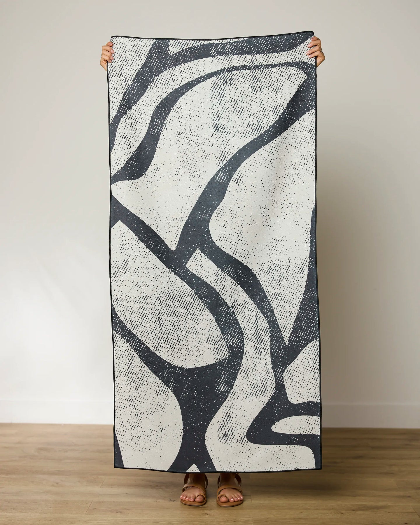 A person holds up the GEOMETRY Block Micro-loop Beach Towel, featuring an abstract black and white rectangular design, hiding their face and body except for hands and brown sandals, set against a plain light background.