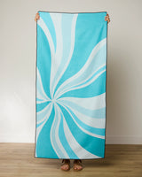 A person stands indoors on a wooden floor, holding up the GEOMETRY Candy Blue beach towel with a swirling white pattern. Only their hands and feet are visible behind this stylish, quick-drying towel.
