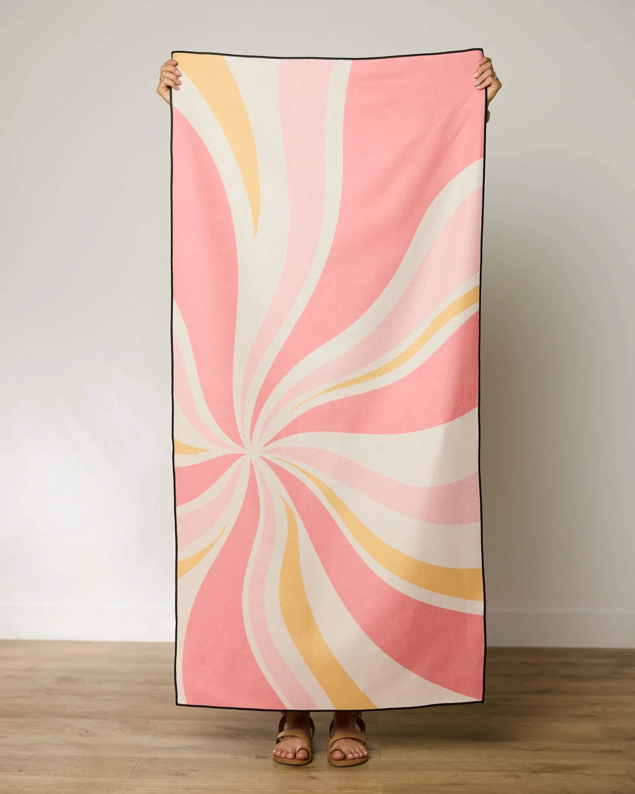 A person stands on a wooden floor, holding up the GEOMETRY Candy Pink Beach Towel with a pink, cream, and yellow swirl design; their face and upper body are hidden behind this quick-drying towel.