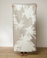 A person holds up the GEOMETRY Sandy Palms Micro-loop Beach Towel, featuring a white palm tree pattern on light beige, standing indoors on a wooden floor with a plain white wall behind them. This quick-drying towel is both stylish and practical.