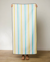 A person stands indoors holding up a GEOMETRY Seaside Sunshine beach towel with vertical blue, yellow, and white stripes; only their hands and sandal-clad feet are visible.