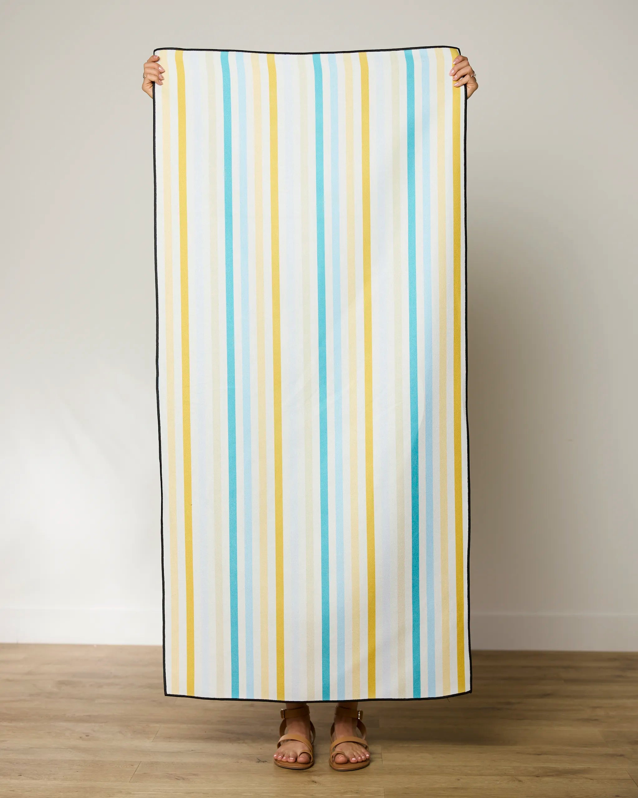 A person stands indoors holding up a GEOMETRY Seaside Sunshine beach towel with vertical blue, yellow, and white stripes; only their hands and sandal-clad feet are visible.