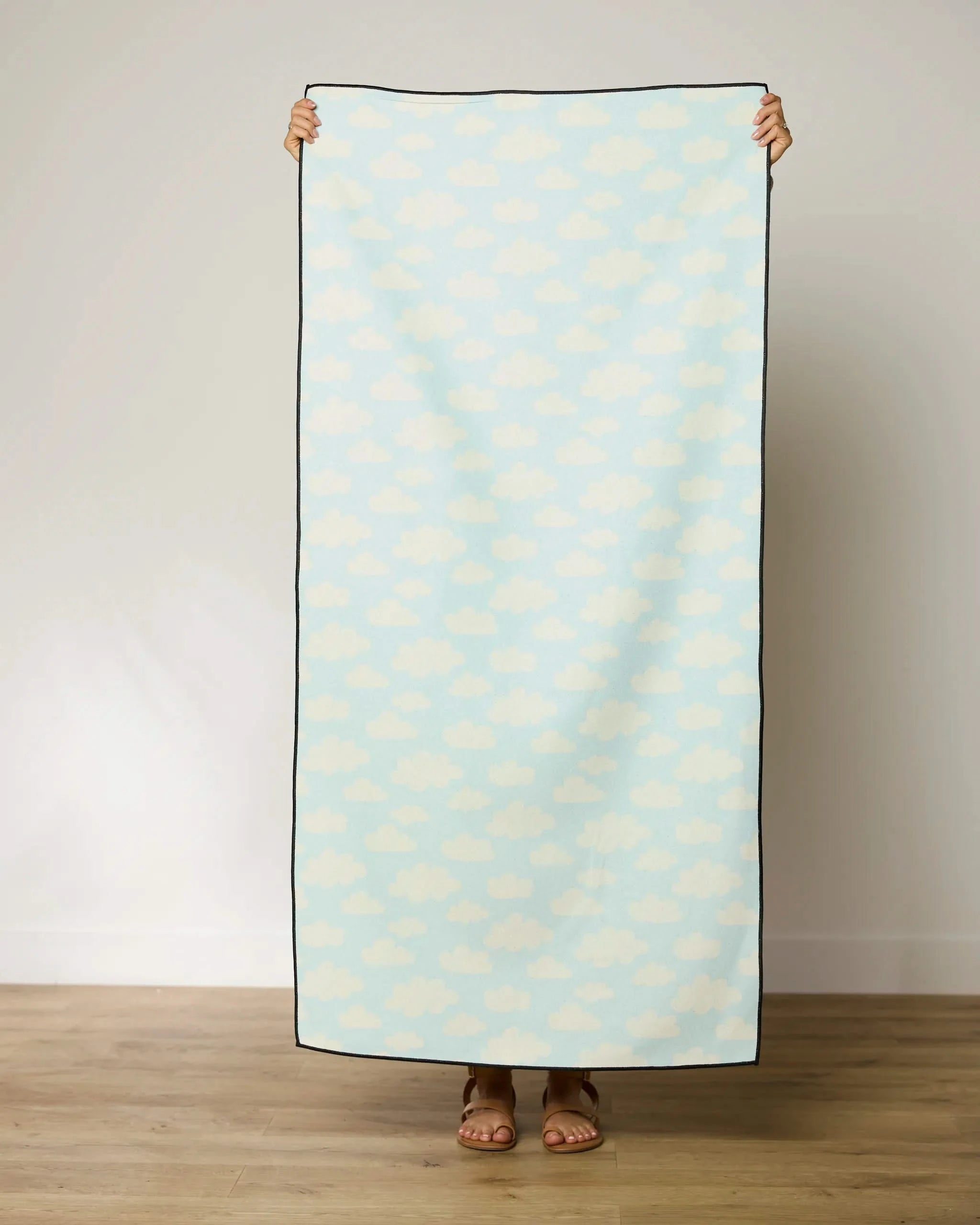 A person stands on a wooden floor holding up the GEOMETRY Made In The Shade beach towel, which has a light blue background with a white cloud pattern, covering their body and showing only hands and feet.