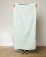 A person stands on a wooden floor holding up the GEOMETRY Made In The Shade beach towel, which has a light blue background with a white cloud pattern, covering their body and showing only hands and feet.