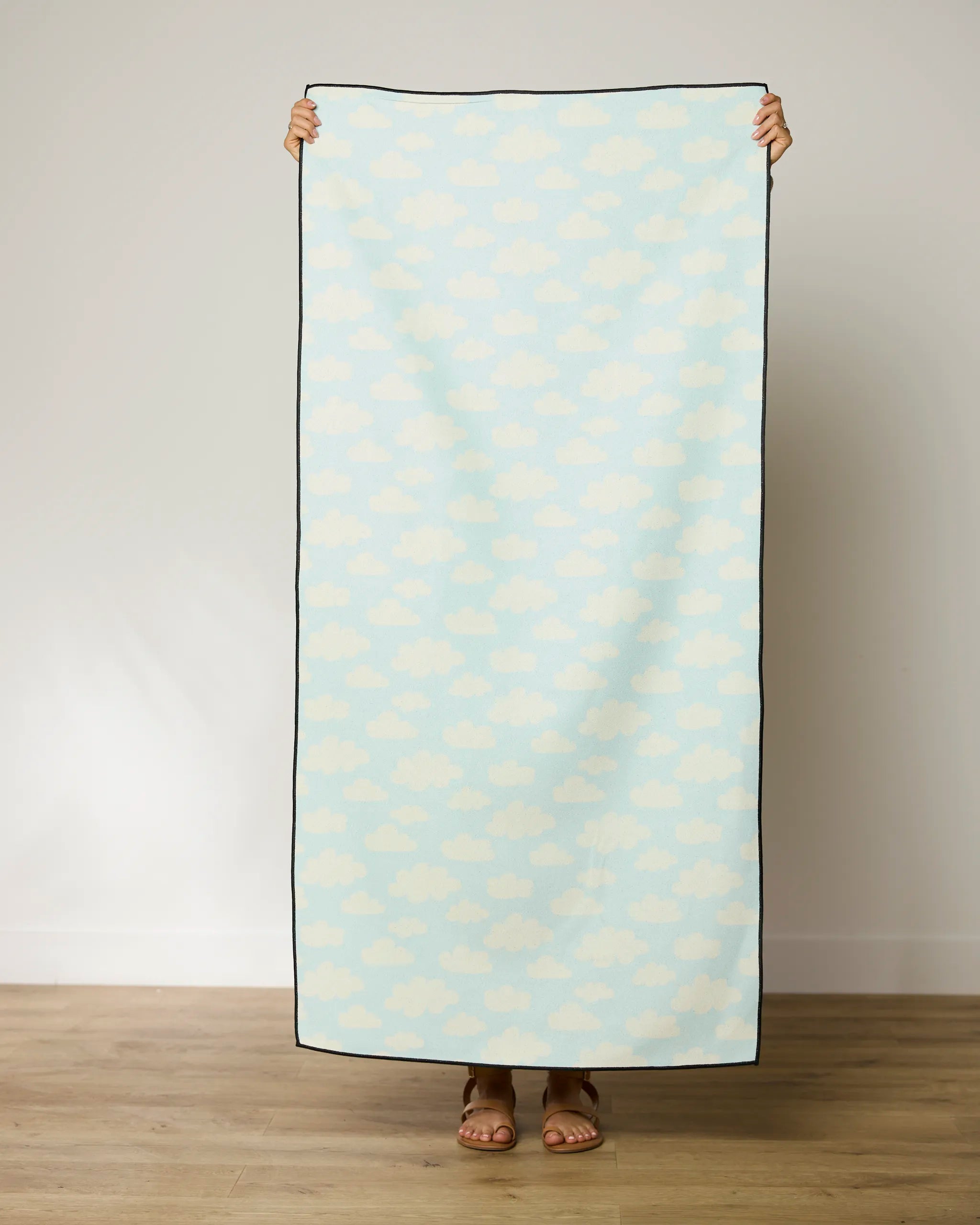 A person stands on a wooden floor holding up the GEOMETRY Made In The Shade beach towel, which has a light blue background with a white cloud pattern, covering their body and showing only hands and feet.