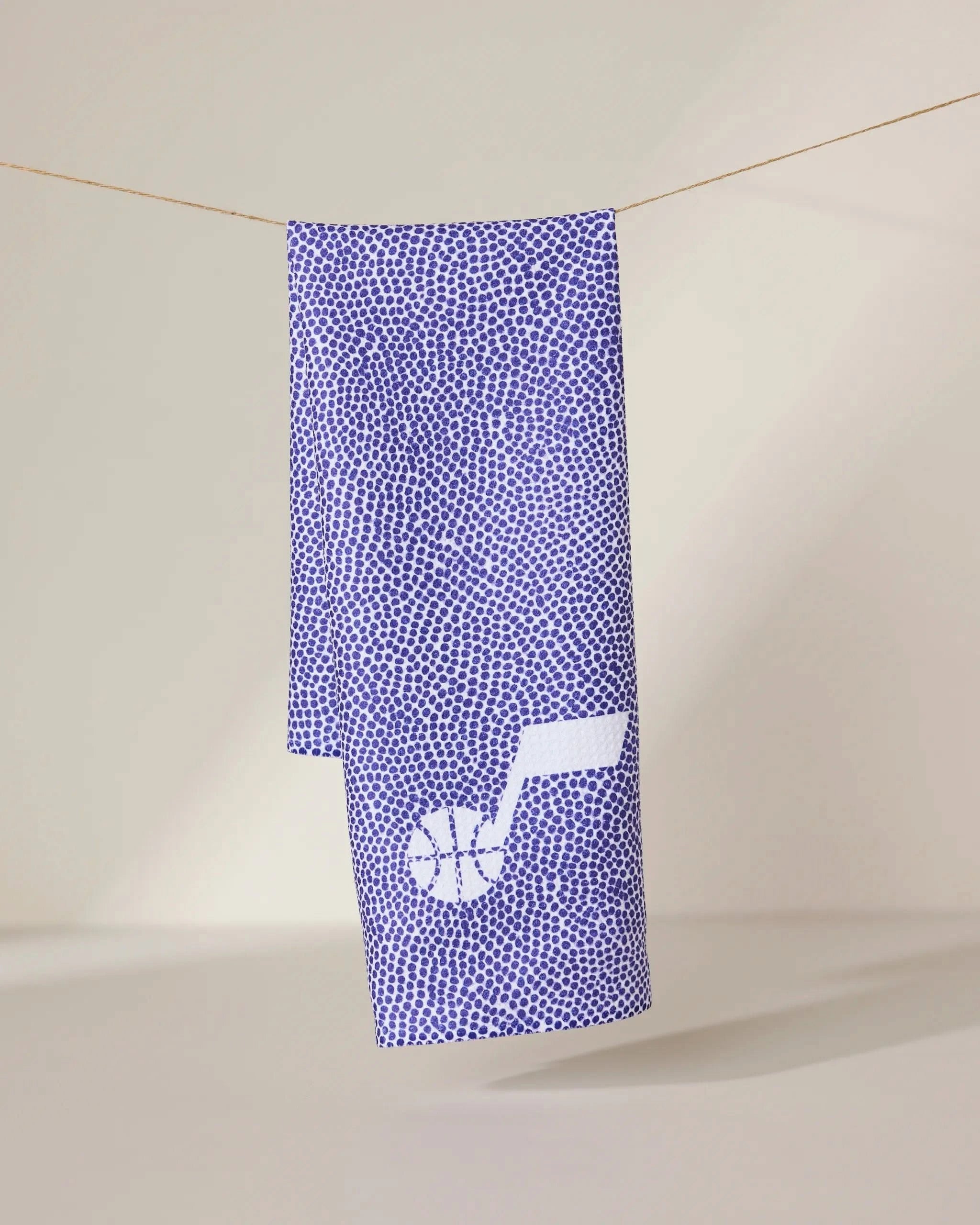The Geometry Jazz Grip towel, featuring a purple and white dotted pattern and a white basketball logo shaped like a musical note in the lower corner, hangs on a softly lit, neutral background.